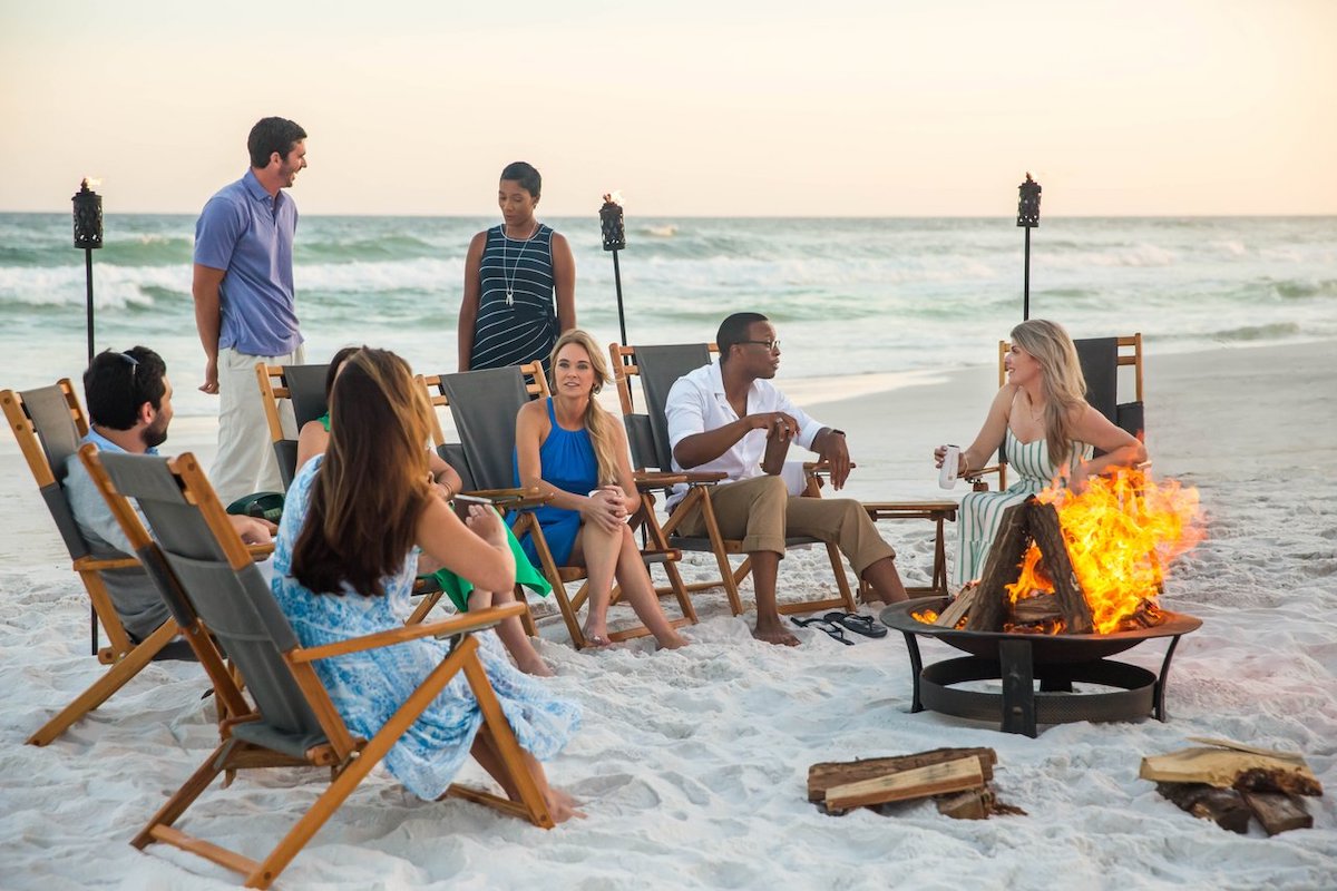 A group of 5 people of mixed genders and races sit around a campfire at dusk on a beach in South Walton, with the ocean visible in the immediate background.